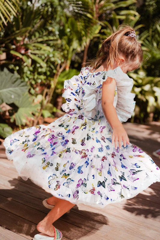 A young girl wearing a white dress with a floral pattern of butterflies in various colors, spinning around in an outdoor setting by Lola and the Boys.