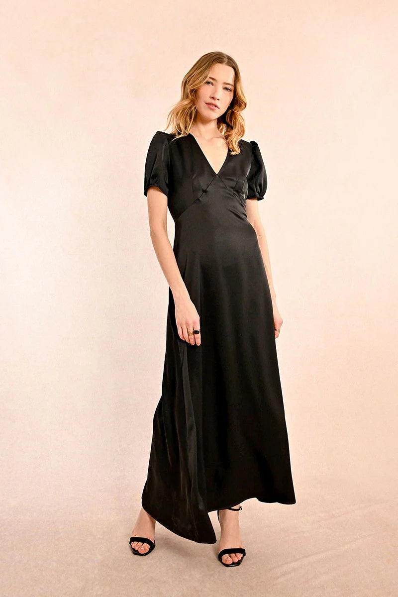 Long black satin dress with short puffed sleeves, a V-neckline, and chest cutouts. The dress has a maxi length and she is paired with black strappy sandals.