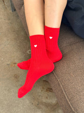 Load image into Gallery viewer, Red socks with heart designs worn on a person's feet on a concrete floor.
