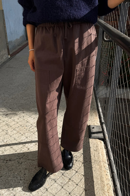 A person wearing brown wide-leg drawstring trousers with a subtle diagonal textured pattern, paired with a dark navy sweater and black loafers, standing outdoors next to a metal fence.