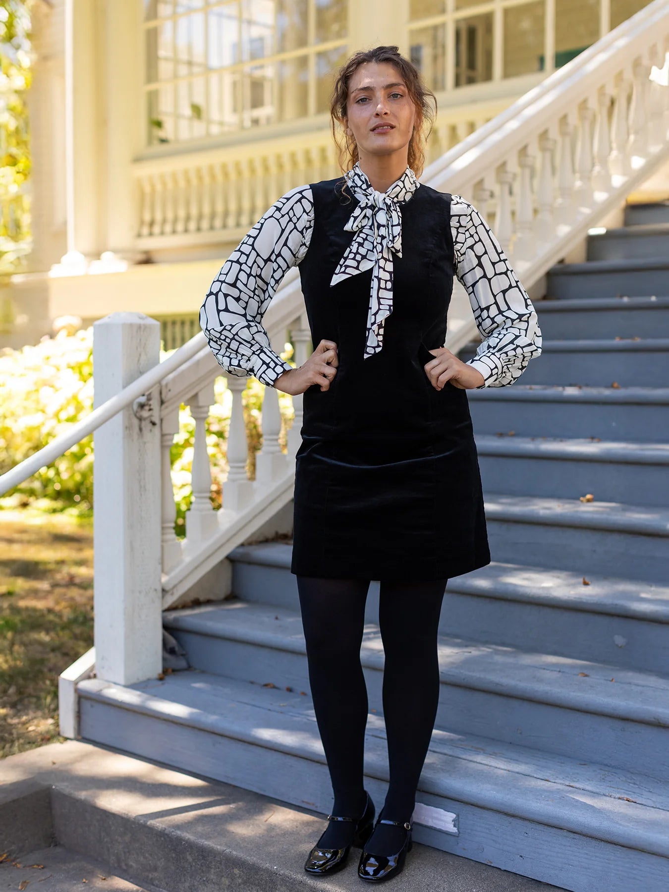 Woman in a black dress with a white patterned blouse standing on steps outdoors.