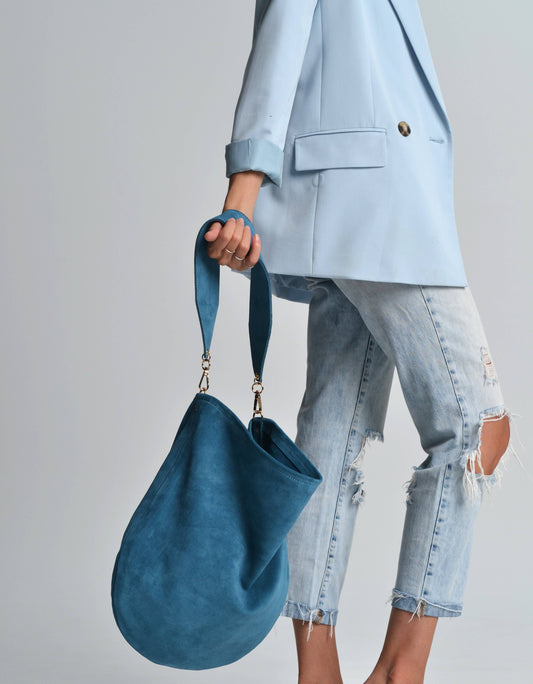 Person holding a blue handbag wearing a light blue blazer and jeans on a gray background